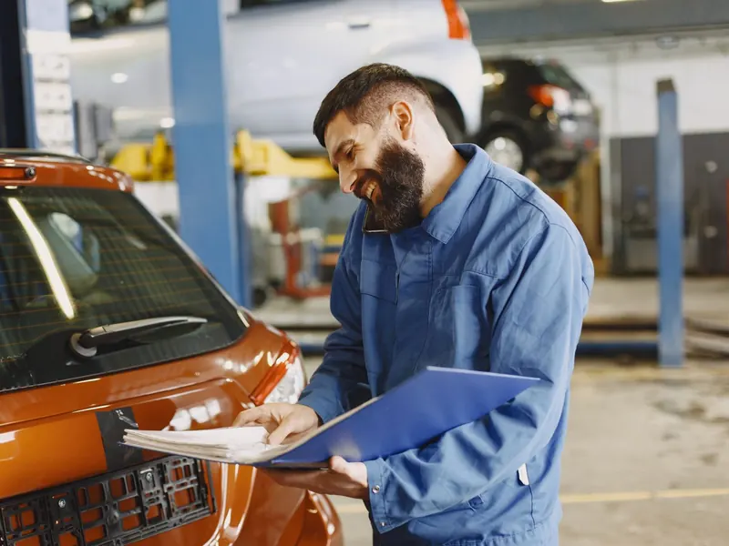 Smiling technician in blue overalls checking a clipboard beside an orange car on a lift