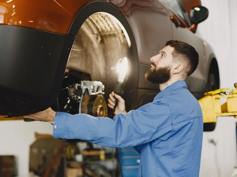 Technician in blue overalls inspecting the brakes behind the wheel of a raised orange car