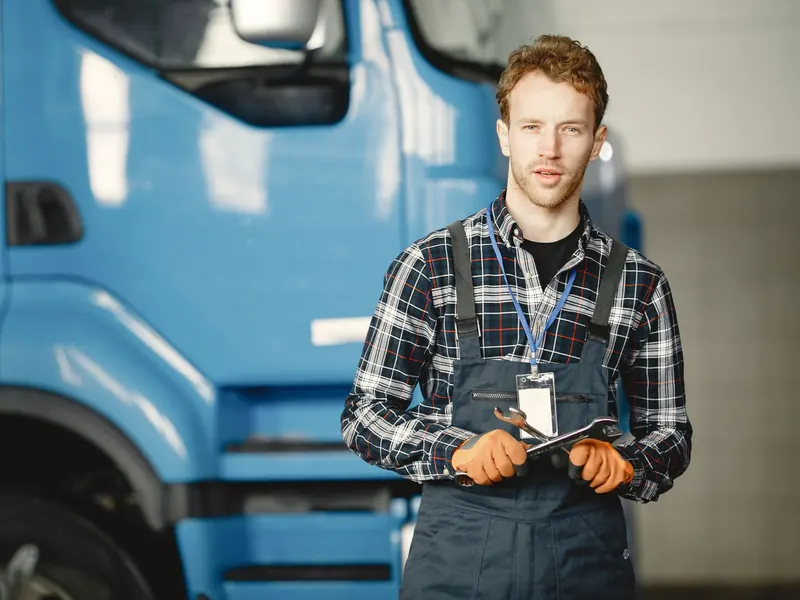 Young technician in plaid shirt and overalls holding a wrench in a truck workshop